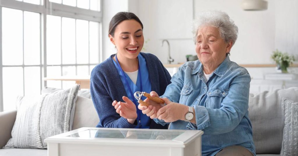 A smiling young female caregiver assisting a happy elderly woman with a hand exercise tool in a bright living room.