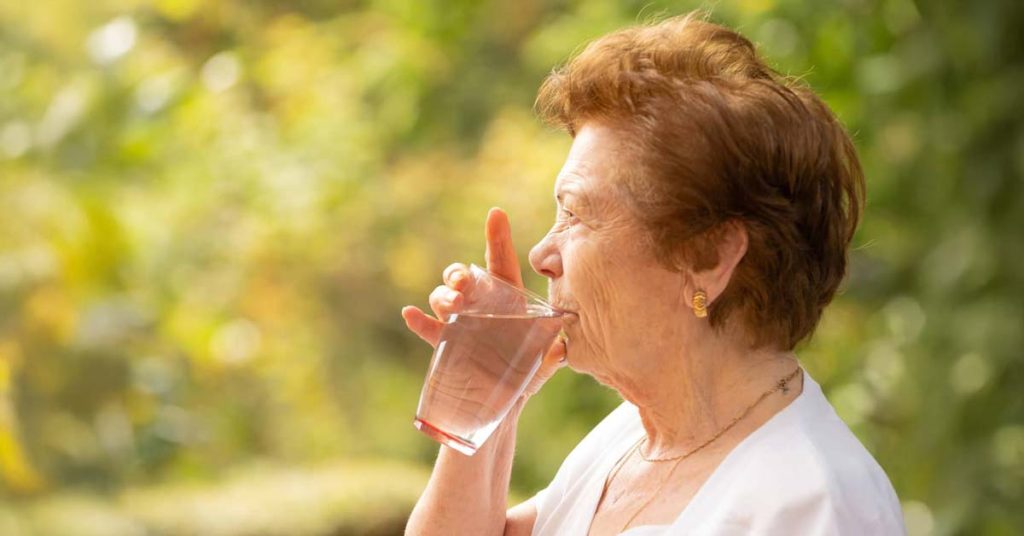 Elderly woman drinking water from a glass outdoors to stay hydrated