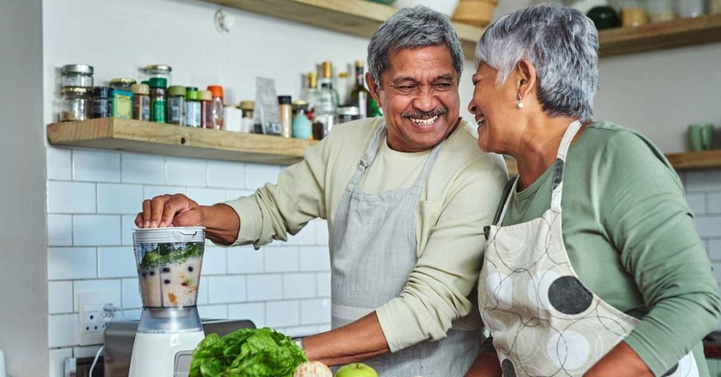 A happy senior couple in a kitchen preparing a healthy fruit and vegetable smoothie in a blender.