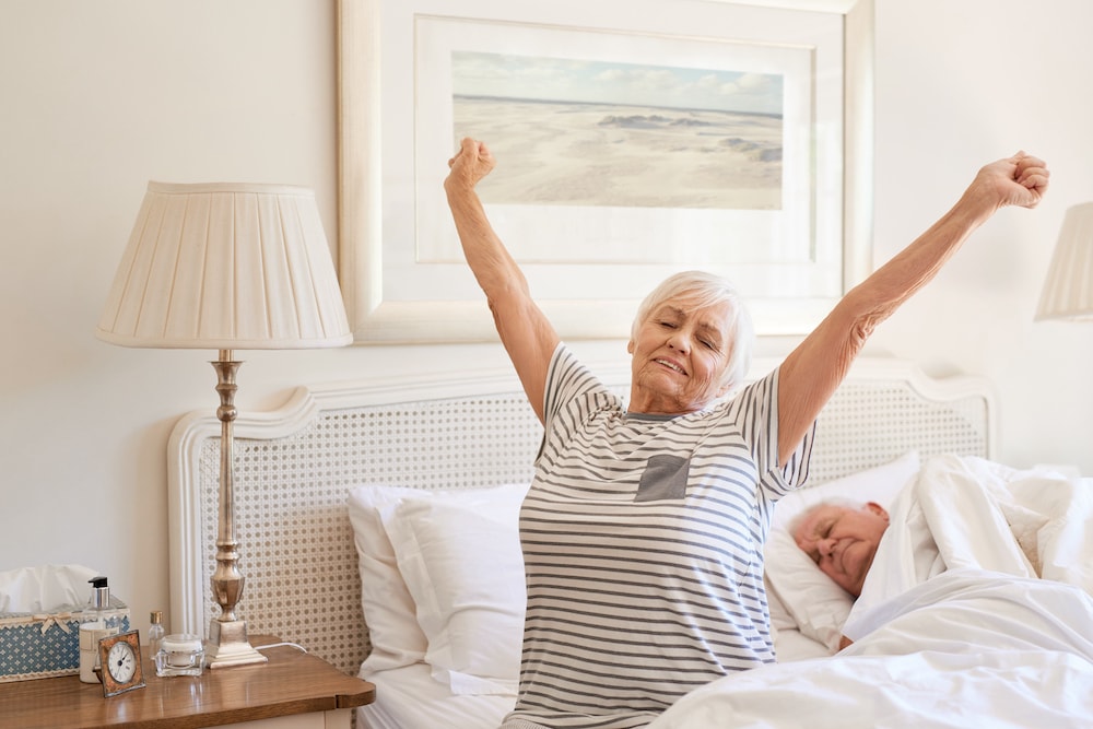 senior woman stretching in bed after waking, improved sleep, elderly couple bedroom morning
