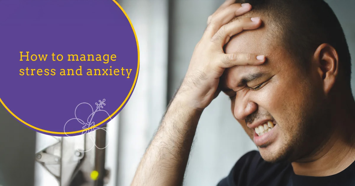An Asian man with a pained expression, his eyes closed and teeth clenched, holds his hand to his forehead as if experiencing a severe headache or intense emotional distress. To his left, a large purple circle outlined in yellow contains the text "How to manage stress and anxiety" in a yellow, serif font. A delicate white outline of a flower is layered over the bottom of the purple circle. The background is softly blurred, showing a window with natural light.