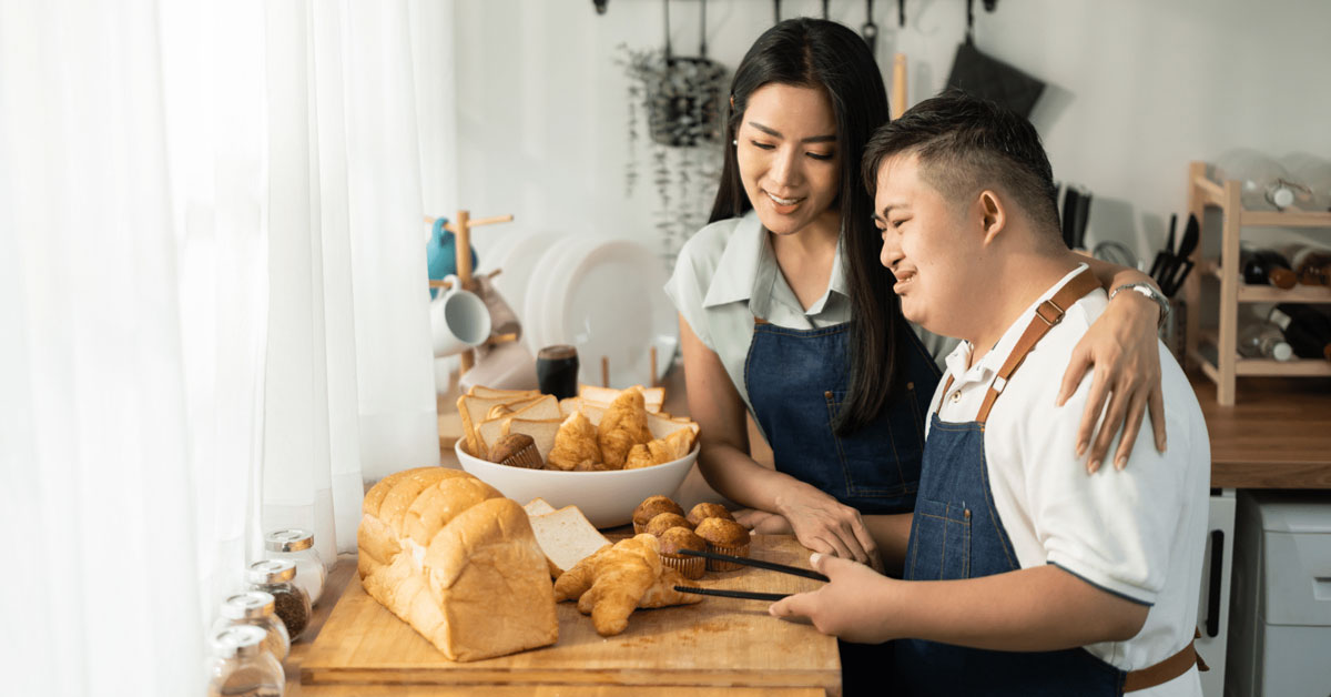 Smiling woman with her arm around a young man with Down syndrome as they happily prepare breakfast together in a bright kitchen – HDE Home Care supports families caring for adults with intellectual and developmental disabilities in Portland, Oregon.