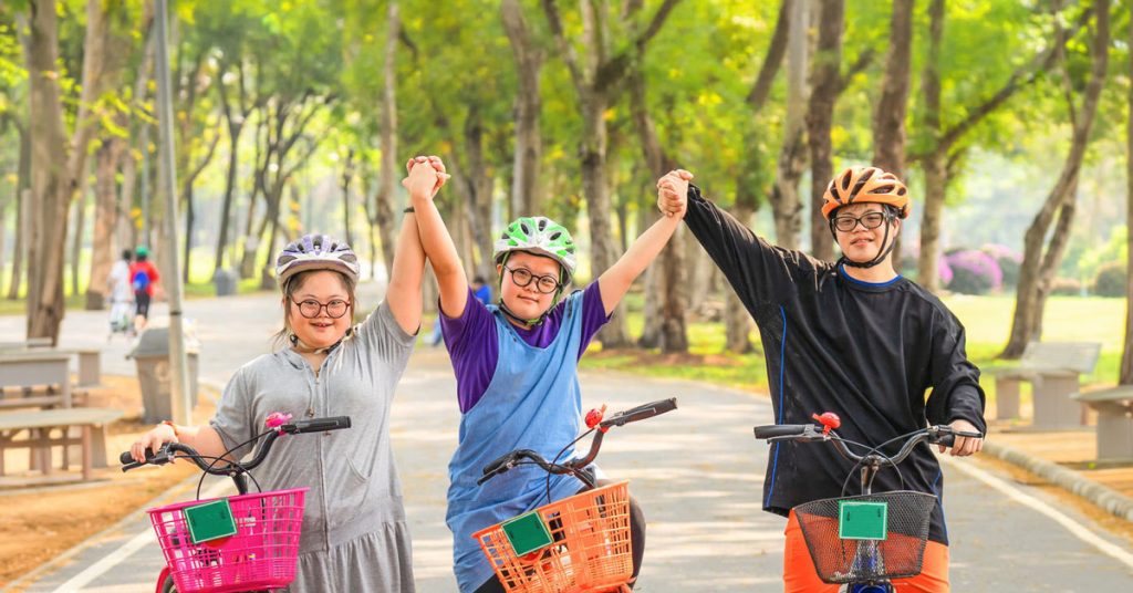 Three smiling youth with Down syndrome holding hands in celebration while riding bicycles on a paved park path.