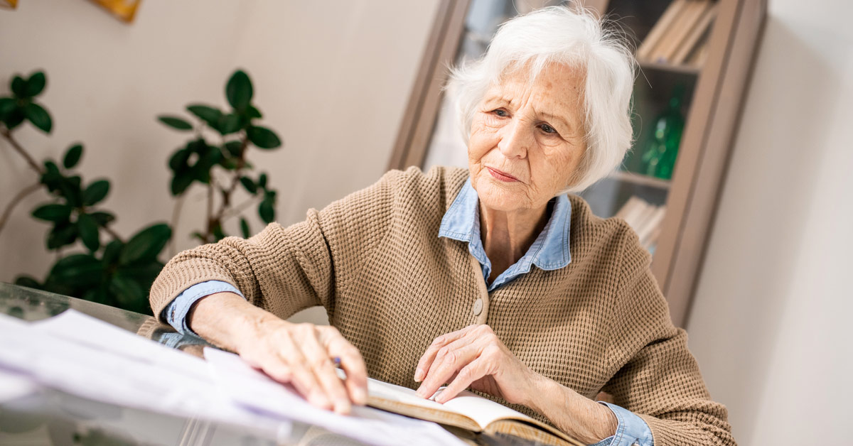 A concerned senior woman with white hair looking down at papers and a book spread across a glass table indoors.