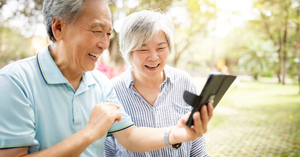 A smiling senior East Asian couple looking at a smartphone together outdoors in a sunny park.