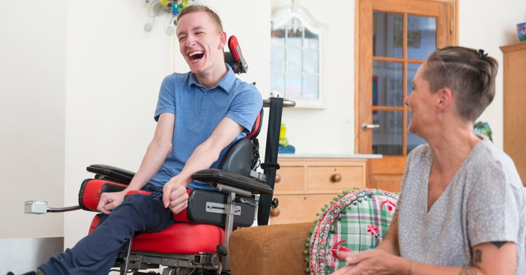 A young man sitting in a specialized motorized wheelchair, laughing heartily during a conversation with a woman sitting opposite him in a bright living room.