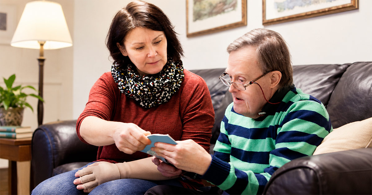 A caregiver sitting on a sofa alongside a man with Down syndrome, both focused on looking through a small booklet or card together in a cozy living room setting.