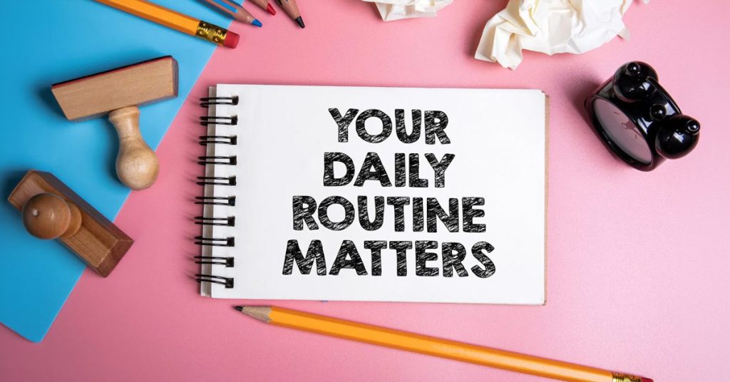 Top view of an open spiral notebook reading "YOUR DAILY ROUTINE MATTERS" in bold text, surrounded by pencils, wooden stamps, crumpled paper, and a small black alarm clock on a pink and blue background.